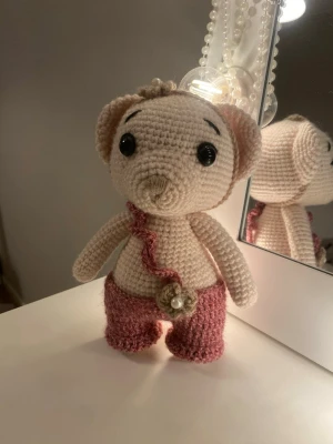 Virkat gosedjur björn med rosa byxor - Handvirkad björn i beige garn med stora svarta ögon och rosa virkade byxor. Dekorerad med en virkad blomma och vågig kant framtill. 