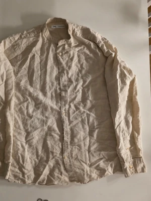 Beige linneskjorta från Zara - Säljer en beige skjorta från Zara i linne med klassisk krage och knappar framtill. Skjortan har lång ärm och en avslappnad passform, perfekt för en chill och stilren look. Enkel att matcha med jeans eller shorts                       Armlängd: 60 cm    Bröstmått: 52.5 cm         Hela: 72 cm