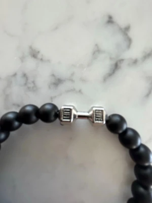 Svart pärlarmband med hantel - Stilrent armband med mattsvarta pärlor och ett coolt silverfärgat hantel-hänge i metall. Perfekt accessoar för dig som gillar gym eller vill ha en sportig touch på din stil. Elastiskt band gör det enkelt att ta på och av.