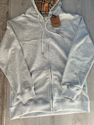 Grå zip hoodie från Burberry - Snygg grå zip hoodie från Burberry med klassiskt rutigt foder i huvan och broderad logga på bröstet. Hoodien har dragsko i huvan, fickor fram och ribbade muddar. Perfekt för dig som vill ha en stilren och exklusiv look.