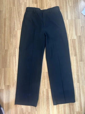 Svarta kostymbyxor med resår - Säljer ett par svarta kostymbyxor med rak passform och resår i midjan. Byxorna har en stilren look och är tillverkade i en mjuk och bekväm mix av rayon, nylon och elastan. Perfekta för dig som gillar en clean och enkel stil.