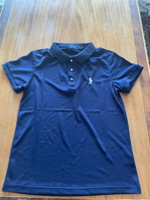 Ralph Lauren piké - Klassisk marinblå pikétröja från Polo Ralph Lauren med korta ärmar, krage och tre knappar framtill. Ikoniska broderade loggan på bröstet. Perfekt till jeans eller shorts för en clean och stilren look.