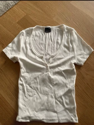 Vit ribbad topp med knappar Gina Tricot - Säljer en vit ribbad topp från Gina Tricot med korta ärmar och djup v-ringning. Toppen har två knappar framtill och är i mjuk bomull. Perfekt till jeans eller kjol för en enkel och fräsch look.
