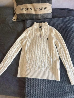 Vit quarter zip tröja Polo Ralph Lauren - Kabelstickad vit tröja från Polo Ralph Lauren med quarter zip och hög krage. Tröjan har det klassiska mörkblå logobroderiet på bröstet och ribbade muddar vid ärmslut och nederkant. Tillverkad i mjukt material, perfekt för sommar kvällar 