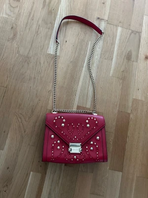Michael Kors Röd axelväska med nitar och kedja - Michael Kors. Snygg röd axelväska i läderimitation med guldfärgad kedjerem och dekorativa nitar framtill. 