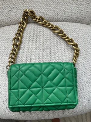 Grön quiltad handväska från Zara - Snygg grön handväska från Zara i quiltat konstläder med en chunky guldfärgad kedja som axelrem. Väskan har en rektangulär form och coolt rutmönster på framsidan. Perfekt för dig som vill sticka ut med en färgglad accessoar.