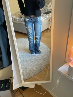 Blå slitna baggy bootcut jeans - Säljer ett par blå bootcut jeans med slitningar och hål på låret. Jeansen har låg midja och klassiska fem fickor. Perfekta för en avslappnad och trendig look. Materialet är denim i bomull.