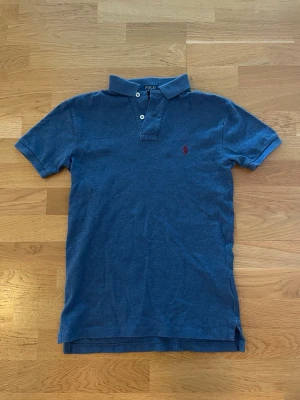 Blå polotopp från Polo Ralph Lauren - Klassisk blå polotopp från Polo Ralph Lauren i slim fit-modell. Tillverkad i mjuk bomull med korta ärmar, krage och knappar framtill. Ikoniska röda loggan broderad på bröstet. Perfekt för en clean och stilren look.