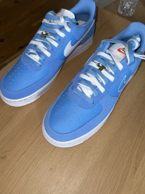 Nike Air Force 1 blå och vita sneakers - Säljer ett par Nike Air Force 1 sneakers i blått med vita detaljer och vit sula. Skorna har klassisk låg siluett, perforerad tåbox och vita skosnören med gulddetaljer. 