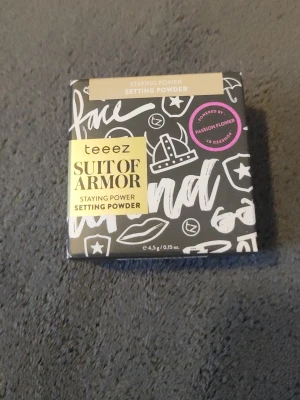 Teeez Suit of Armor Setting Powder - Teeez Suit of Armor Setting Powder är ett kompakt puder i en svart och vit förpackning med guldiga detaljer. Pudret är designat för att fixera foundation och ge en matt finish. Förpackningen innehåller 4,5 g och är dekorerad med grafiska mönster och text.