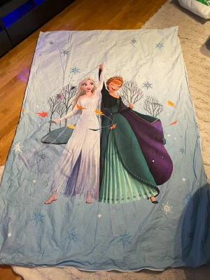 Frost påslakan med Elsa och Anna 140cm brett - Säljer ett blått påslakan med motiv av Elsa och Anna från Disney-filmen Frost. Ena sidan har en stor bild av karaktärerna och snöflingor, andra sidan har ett mönster med små granar och blommor. Materialet är mjukt bomullstyg, perfekt för dig som gillar Frost.