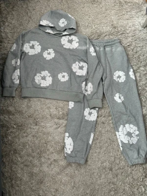 Grått denim tears set - Mysigt denim tears set i grått med vita blom tryck. Setet består av en hoodie med känguruficka och matchande byxor med resår i midja och benslut. Perfekt för chill dagar hemma eller när du vill vara extra bekväm.