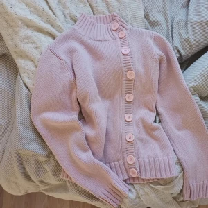 Ljusrosa stickad kofta med knappar - Säljer en söt ljusrosa stickad kofta med stora matchande knappar framtill. Koftan har ribbade muddar vid ärmslut, nederkant och hals, samt en högre krage. Perfekt för lager på lager och ger en mysig vibe till din outfit.