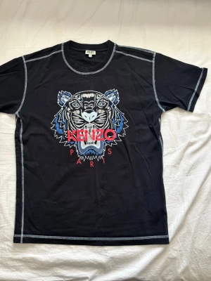 Svart Kenzo t-shirt med tigertryck - Svart t-shirt från Kenzo med ikoniskt tigertryck i blått, vitt och beige samt röd Kenzo Paris-text på bröstet. T-shirten har kontrastsömmar och rund hals. Perfekt för dig som vill sticka ut med en streetstyle-look.