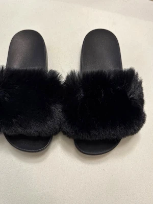 Svarta fluffiga slip-in tofflor - Säljer ett par svarta slip-in tofflor med bred rem i mjuk, fluffig fuskpäls. Sulan är i gummi med räfflad undersida för bättre grepp. Perfekta för dig som vill ha något bekvämt och trendigt hemma eller till vardagslooken.