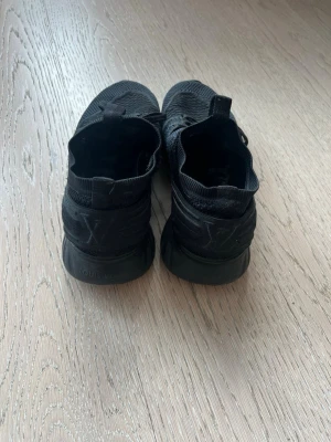 Svarta Louis Vuitton sneakers - Snygga helsvarta sneakers från Louis Vuitton med stickad ovandel och diskret LV-logga på hälen. Skorna har en sportig siluett, snörning och tjock sula för extra komfort. Perfekta för dig som gillar exklusiva och stilrena sneakers.