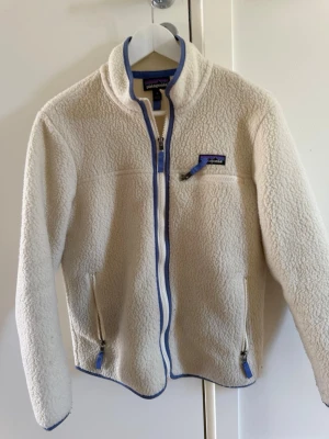 Vit fleecejacka från Patagonia - Mysig vit fleecejacka från Patagonia med blå detaljer vid dragkedjor och kanter. Jackan har hög krage, hel dragkedja framtill och tre fickor med dragkedja. Perfekt för lager-på-lager och outdoor-vibe. 