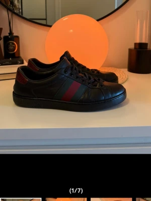 Svarta Gucci sneakers  - Säljer ett par svarta skor från Gucci i läder med den ikoniska röd och grön randen på sidan. Skorna har lågt skaft, svarta skosnören och en röd detalj vid hälen. Perfekt för dig som gillar exklusiv och stilren design.