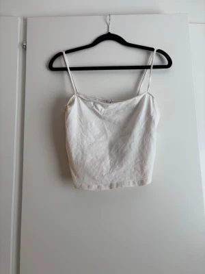 Vit croppad linnetopp - Säljer en enkel och stilren vit croppad linnetopp med tunna axelband. Perfekt att matcha med jeans eller kjol för en fräsch look. Toppen har en rak passform och är ärmlös.