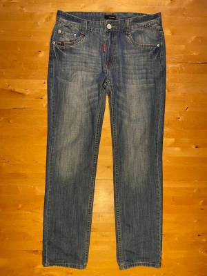 Blå raka jeans från Diesel - Säljer ett par klassiska blå jeans från Diesel med raka ben och fem fickor. Jeansen har subtila slitningar och röda detaljer vid fickorna. Perfekta för en avslappnad och stilren look.