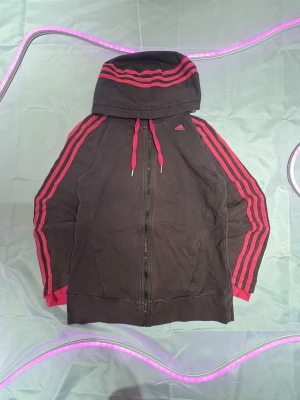 Svart hoodie med rosa ränder Adidas - Säljer en svart hoodie från Adidas med dragkedja och huva. Hoodien har ikoniska rosa ränder längs ärmarna och huvan samt rosa snören. Adidas-loggan syns på bröstet. Perfekt för en sportig och avslappnad stil.