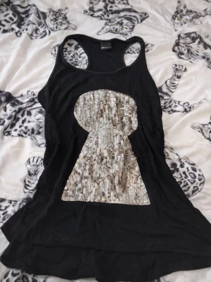 Svart linne med paljettmotiv - Snyggt svart linne med racerback och stort nyckelhålsmotiv i silverfärgade paljetter på framsidan. Tillverkat i ett mjukt och lätt tyg som faller snyggt. Perfekt för dig som vill sticka ut lite extra.