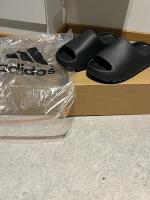 Svarta Adidas Yeezy Slides sandaler - Svarta Adidas Yeezy Slides med chunky sula och öppen tå. Tillverkade i ett mjukt och flexibelt syntetmaterial som ger en clean och modern look. Perfekta för chill dagar och enkel att matcha med streetwear. Levereras med originalpåse.