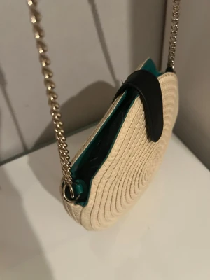 Rund axelväska i strå med kedjerem - Säljer en rund axelväska i naturfärgat stråmaterial med guldfärgad kedjerem och svart läderdetalj på locket. Väskan har en unik cirkelform och insida i grönt tyg. Perfekt för dig som vill ha en trendig accessoar med boho-vibe.