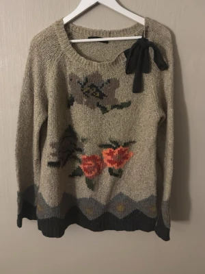 Beige stickad tröja med blommor Twin Set - Beige stickad tröja från Twin Set Simona Barbieri med stora blommor i rosa, rött och grått framtill. Tröjan har en svart rosett vid halsringningen och mörkare ribbstickade muddar samt nederkant. Perfekt för dig som gillar unika detaljer och mjuka material.