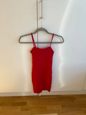 Röd kort klänning med smala axelband - Säljer en röd kort klänning med smala axelband och enkel, figurnära passform. Perfekt för dig som gillar minimalistisk stil och vill ha en klänning som sticker ut med sin starka färg. Materialet känns mjukt och stretchigt.