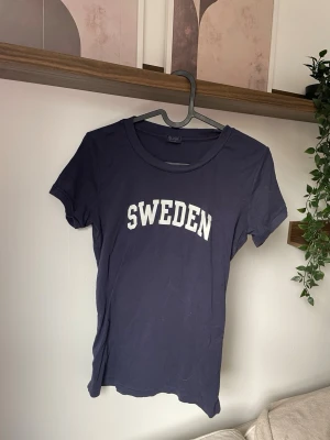 Mörkblå t-shirt med SWEDEN-tryck - Snygg mörkblå t-shirt med vit SWEDEN-text på bröstet. Klassisk rund halsringning och korta ärmar. Perfekt för dig som vill visa lite svensk stolthet på ett stilrent sätt.