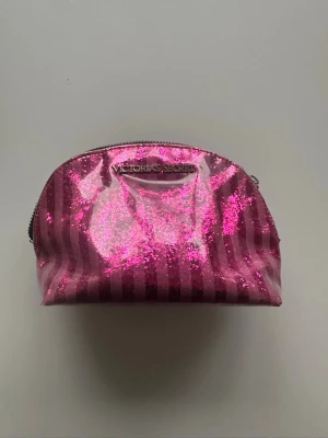 Glittrig rosa necessär Victoria's Secret - Säljer en glittrig rosa necessär från Victoria's Secret med metallicrandigt mönster och dragkedja. Väskan har en halvmåneform och är täckt av rosa glitter, perfekt för smink eller småprylar. Märket syns tydligt framtill i metall. Tyvärr saknas lilla piggen till dragkedjan men det går att stänga och öppna nessären. Längd ca 21cm höjd 14cm bred 8cm