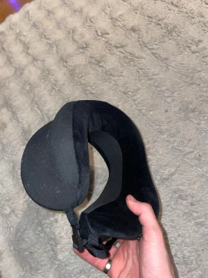 Svart resenackkudde med spänne - En svart U-formad resenackkudde i mjukt tyg, perfekt för att vila huvudet under resor. Kudden har ett justerbart spänne framtill för extra stöd och enkel fastsättning på väskan. Enkel att ta med och superskön för långa resor.