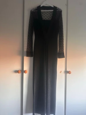 Svart glittrig jumpsuit med spetsärmar - Snygg svart jumpsuit med långa, transparenta spetsärmar och glittrigt tyg. Djup v-ringning och vida ben ger en trendig siluett. Materialet är stretchigt och bekvämt.