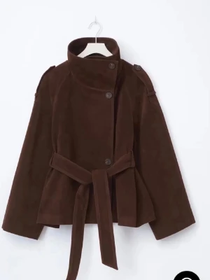 Mörkbrun trenchcoat med bälte - Stilren mörkbrun trenchcoat i mjukt mockaimitationsmaterial. Jackan har hög krage, knäppning med stora knappar och axelklaffar. Midjebälte för snygg passform och vida ärmar ger en modern look. Perfekt för dig som vill ha en trendig och tidlös jacka.