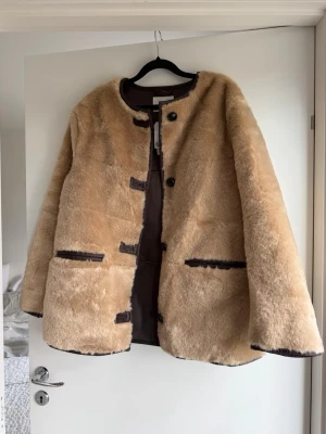 Beige teddyjacka med bruna detaljer - Mysig beige teddyjacka med rund halsringning och knäppning framtill. Jackan har två stora fickor och bruna detaljer längs kanterna och vid knapparna. Oanvänd 