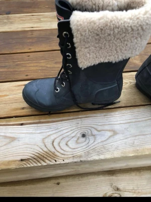 Svarta Hunter vinterstövlar med foder - Säljer ett par svarta snörstövlar från Hunter med högt skaft och tjockt beige teddyfoder. Stövlarna har robust gummiyta och klassisk Hunter-logga framtill. Perfekta för kalla dagar när du vill hålla både stil och värme.