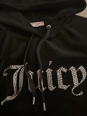 Svart Juicy Couture hoodie med strass - ‼️Mycket mindre i storleken !‼️ Sitter som storlek S. Snygg svart hoodie från Juicy Couture i mjuk velour med huva. Framsidan pryds av texten 'Juicy' i stora bokstäver av glittrande strass. Perfekt för dig som gillar en lyxig look. PRIS KAN DISKUTERAS 
