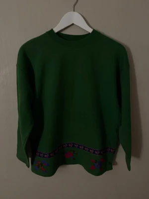 Grön tröja med broderade blommor från United Colors of Benetton. - Grön långärmad tröja med rund hals och broderade blommor i rött, lila och blått längs nederkanten. Dekorativt band med blommönster ovanför broderiet. Perfekt för dig som gillar färgglada detaljer och unik stil.