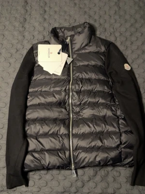 Svart Cardiganjacka från Moncler - Snygg svart pufferjacka från Moncler med quiltad front och hög krage. Västen har stickade svarta ärmar och ett Moncler-märke på ena ärmen. Dragkedja framtill och klassisk design som är både stilren och modern. Perfekt för lager-på-lager och att hålla sig varm. STORLK PASSAR S, HELT NY OCH OANVÄND 