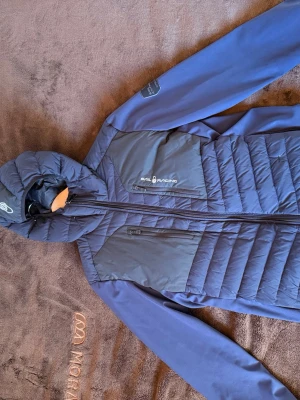 Mörkblå pufferjacka från Sail Racing - Snygg mörkblå pufferjacka från Sail Racing med huva och dragkedja framtill. Jackan har två bröstfickor med dragkedja och quiltad design på framsidan. Perfekt för kalla vinterdagar och riktigt stilren look.