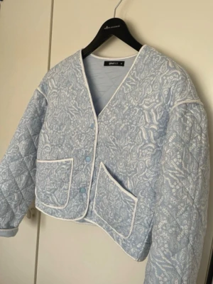 Ljusblå quiltad cardiganjacka Gina Tricot - Supersöt ljusblå quiltad cardiganjacka från Gina Tricot med vitt blommönster och vita kanter. Jackan har två stora fickor framtill, tryckknappar och en croppad passform. Perfekt för dig som vill ha en trendig och mysig look.