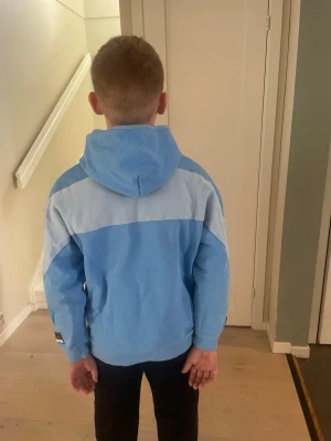 Nike air blå hoodie med huva - Säljer en ljusblå hoodie från Nike med vit logga på bröstet och en bredare ljusare panel över axlarna. Hoodien har en stor huva och ribbade muddar vid ärmslut och nederkant. Perfekt för chill dagar eller när du vill ha en sportig look.