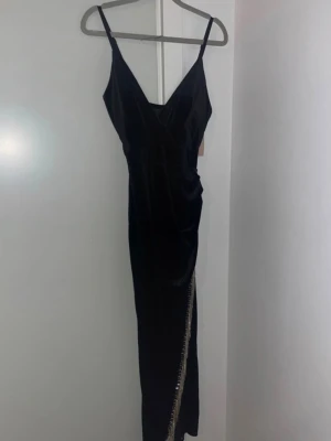 Svart långklänning med fransar - Snygg svart långklänning med smala axelband och v-ringning. Klänningen har en slits framtill dekorerad med glittriga fransar som ger extra wow-känsla. Perfekt för dig som vill sticka ut på festen.