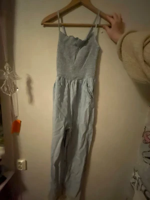 Ljusblå smockad jumpsuit med smala band 158/164 - Supersöt ljusblå jumpsuit med smala axelband och smockad överdel. Byxdelen är lös och luftig, perfekt för sommardagar. Materialet känns mjukt och bekvämt bomull. Enkel att styla med sneakers eller sandaler.