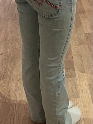 Ljusblå bootcut jeans med låg midja - Säljer ett par lågmidjade ljusblå bootcut jeans med hål vid knäna och rosa broderi på bakfickan. Jeansen har klassisk femficksdesign. Jag har sytt upp dem då de var lite långa men kan såklart ta bort det om man vill. De är från Nelly i storlek 32.