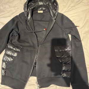 Svart pufferjacka med huva - Snygg svart pufferjacka från Moncler med glansiga quiltade partier på huva, sidor och rygg. Jackan har dragkedja framtill, två fickor och justerbar huva med snören. Materialmix av blankt nylon och mjuk bomull ger en modern look. Ena fickan har släppt lite men går att åtgärda enkelt!