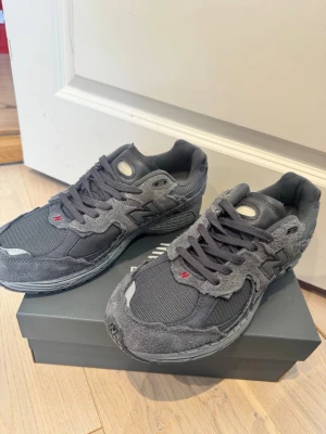 Grå New Balance 2002R sneakers - Säljer ett par grå New Balance 2002R sneakers med slitna mockadetaljer och meshpaneler. Skorna har en chunky sula och klassisk N-logga på sidan. Snygga detaljer i olika grå nyanser och små röda accenter på tungan. Perfekta för en streetwear-look.