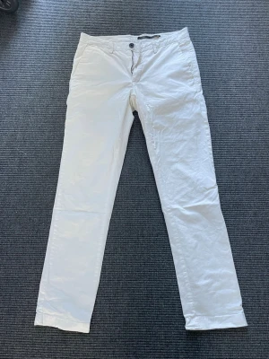 Vita chinos Doppelgänger - Snygga vita chinos med rak passform och klassisk design. Byxorna har framfickor, bakfickor med knapp och stängs med knapp och dragkedja. Perfekta för en clean och stilren look. Materialet känns lätt och passar bra till många outfits.