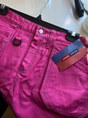 Rosa arbets kjol från Fristads - Cool rosa arbets kjol från Fristads med flera stora fickor och praktiska detaljer som D-ring och reflexband. Kjolen har en kort passform och är gjord i slitstark bomull, perfekt för dig som gillar streetwear och funktionella plagg.